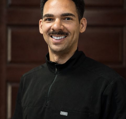 Dr. Josh Thomas smiling in black jacket against wood paneled office background